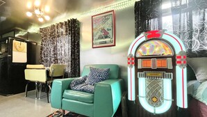 Una sala de estar de estilo retro con un reproductor de discos, un sofá color turquesa, cortinas estampadas y un cartel enmarcado en la pared.