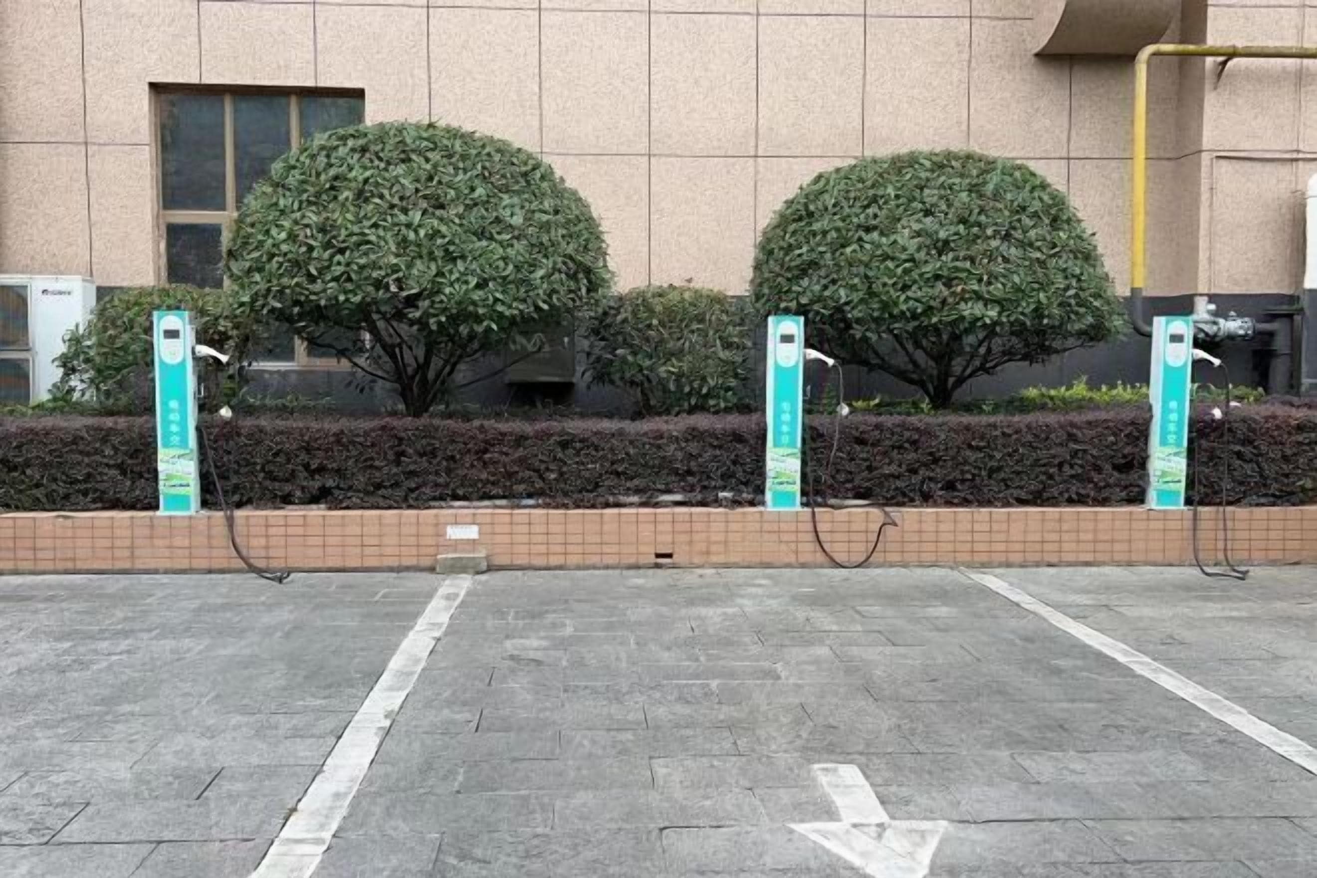 A parking area with electric vehicle charging stations, neatly arranged bushes, and a building with a tiled facade in the background.