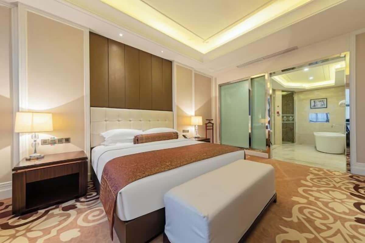 Quarto de hotel com uma cama grande, uma mesa de cabeceira, um abajur, uma porta de vidro que dá para um banheiro e um tapete estampado.