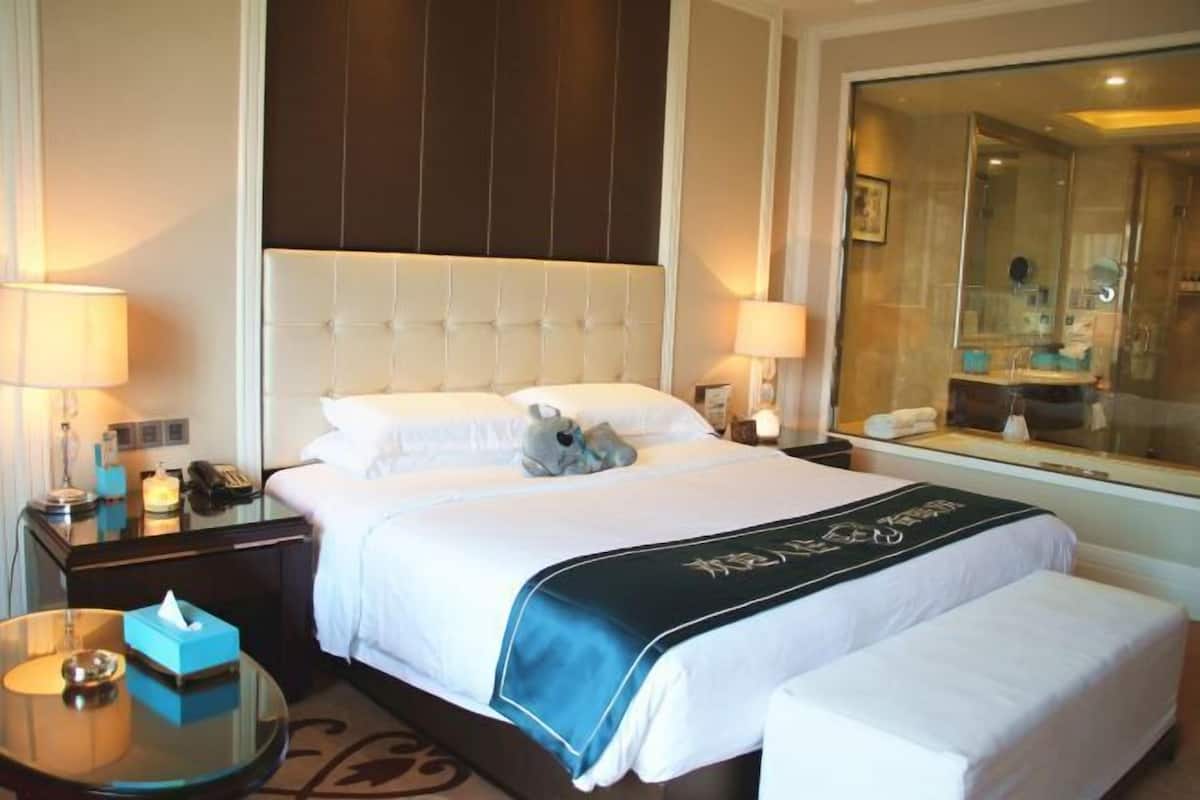 Quarto de hotel com uma cama grande, mesas de cabeceira, uma mesa de centro de vidro, um espelho e um banheiro visível pela porta de vidro.
