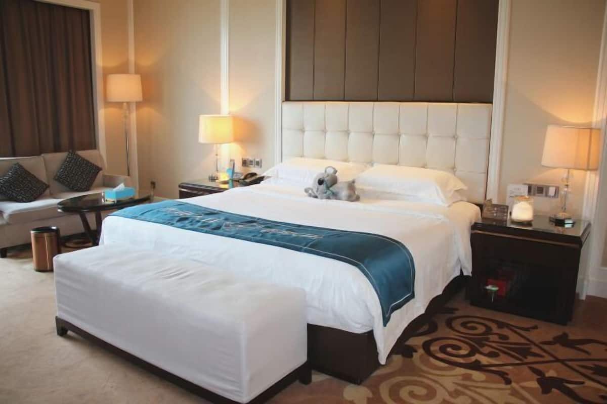 Quarto de hotel com uma cama grande, mesas de cabeceira, luminária, sofá e tapete estampado.
