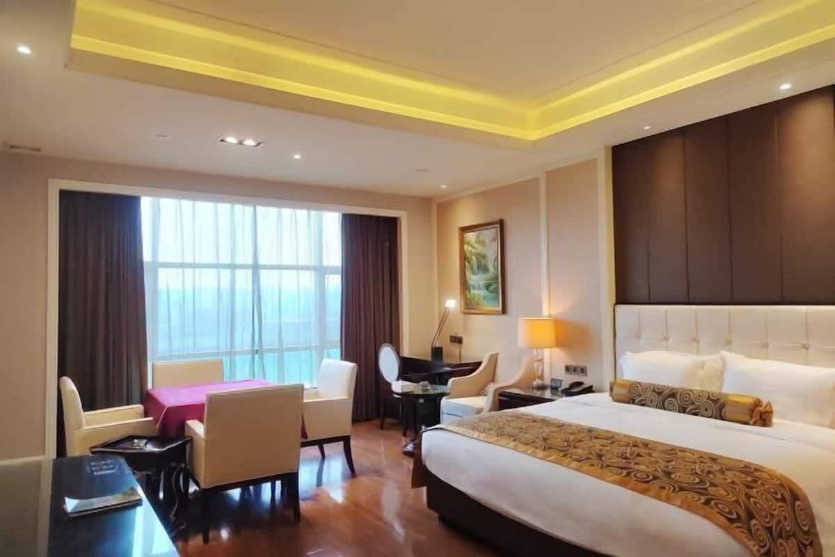 Quarto de hotel com uma cama grande, uma escrivaninha com cadeira, uma mesinha e uma janela com cortinas.