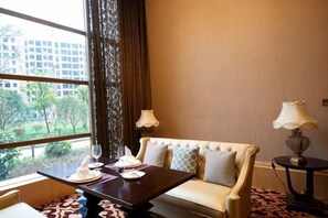 Ein Hotelzimmer mit einer Couch, einem Esstisch mit Tellern und Gläsern, zwei Lampen und Blick auf Gebäude und Bäume durch das Fenster.