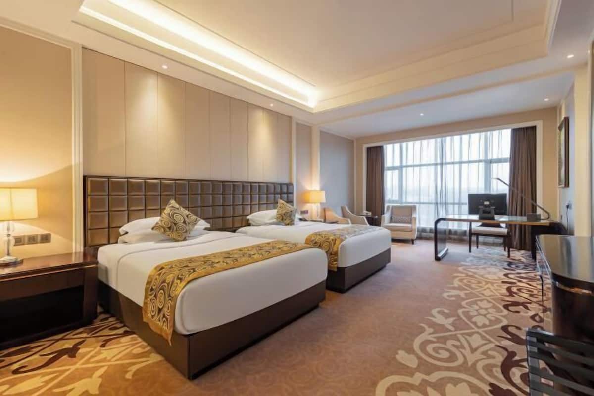 Quarto de hotel com duas camas, uma mesa, uma cadeira, uma televisão e uma janela grande.