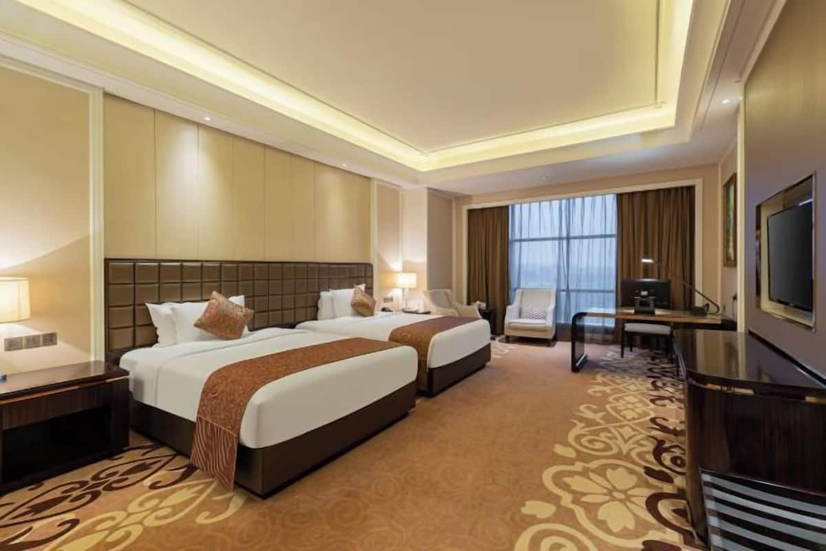 Quarto de hotel com duas camas, uma escrivaninha, uma televisão e uma janela grande.