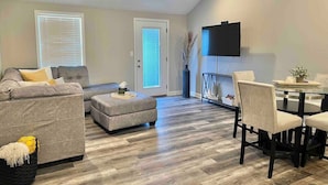A living room with a gray sofa, an ottoman, a dining table with chairs, a flat-screen TV, and a ceiling fan. - Modern 2bd 2ba - Near I75, No Steps, Free Wifi (Richmond)