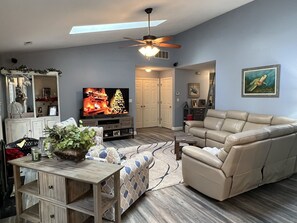A living room with a beige sofa, a TV on a wooden stand, a ceiling fan, and a large window.