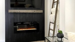 A modern living room with a black wall-mounted TV, a fireplace, a glass coffee table, and a ladder leaning against the wall. - Modern Getaway in Downtown Bozeman! (Bozeman)