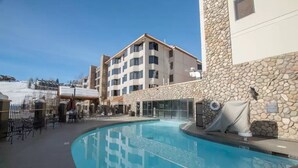 A swimming pool area with a stone wall and a building with multiple balconies. - Lift Base Village - Grand Lodge King Bedroom Suite (Mount Crested Butte)