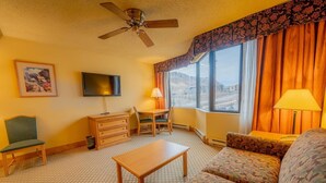 A hotel room with a ceiling fan, a flat-screen TV, a wooden dresser, a dining table with chairs, a sofa, and large windows with curtains.