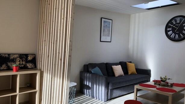 A modern living room with a grey sofa, a wooden partition, a framed picture, a black clock, and a small table with red cushions. - Charming apartment, Left bank (Rouen)