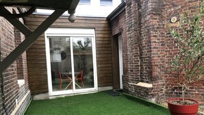 A small backyard with artificial grass, a red potted plant, and a sliding glass door leading to a patio area with a table and chairs. - Charming apartment, Left bank (Rouen)