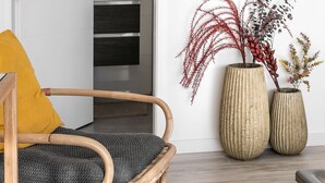 Una habitación con una silla de mimbre, un cojín amarillo y dos jarrones grandes con plantas secas sobre un piso de madera.