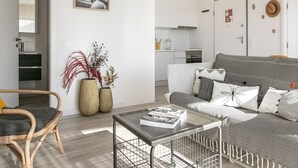 Una sala moderna con un sofá, una mesa de centro y una silla de mimbre. Se observa una cocina al fondo con armarios blancos y una estufa.