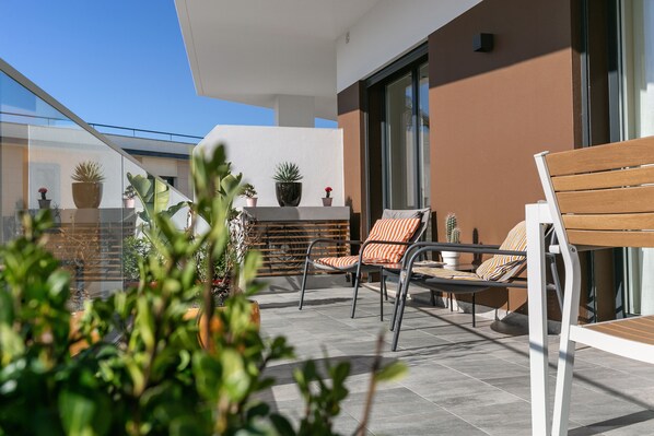 A balcony with two chairs and a table, potted plants, and a modern building facade.