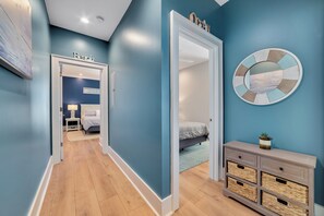 Un corridoio che porta a una camera da letto con un letto, un comodino e una lampada. Si nota uno specchio fissato al muro e un cartello decorativo sopra il letto.