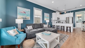 A living room with a grey sofa, a blue armchair, and a white coffee table. There is a kitchen area with white cabinets and a dining set.