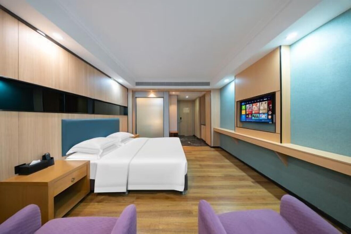 Une chambre d’hôtel avec un lit, un téléviseur fixé au mur, une table de chevet en bois et un fauteuil violet.