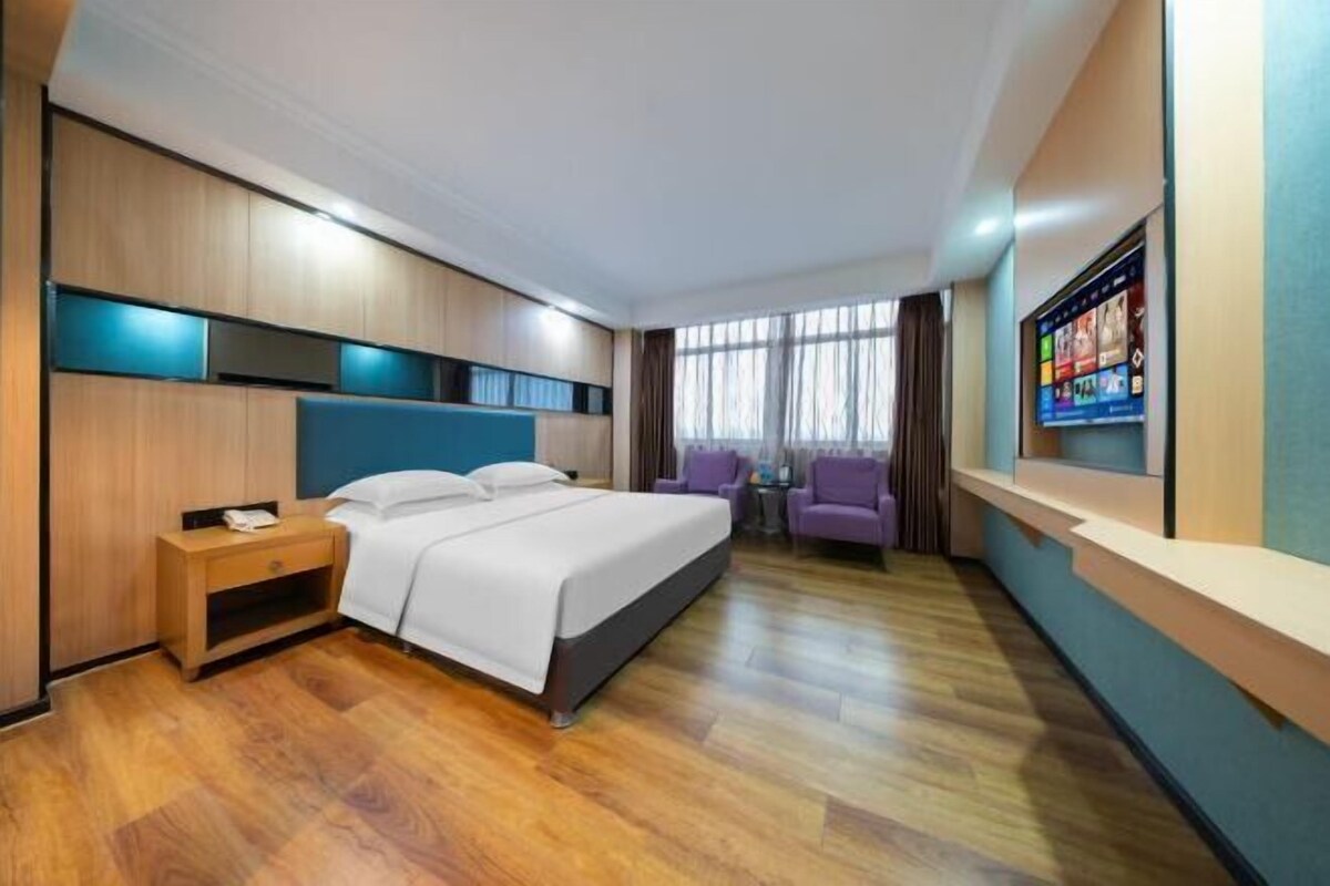 A hotel room with a bed, bedside table, two chairs, a TV, and a window with curtains.
