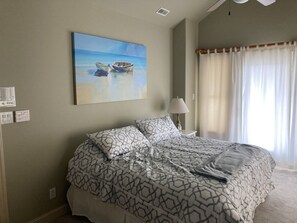 A bedroom with a bed, a painting of boats on a beach, a lamp, and a window with curtains.