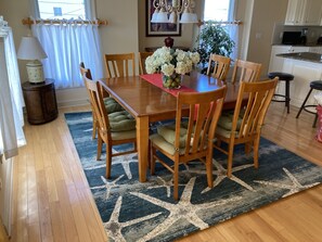 A dining area with a wooden table, six chairs with cushioned seats, a vase of white flowers, and a blue patterned rug.