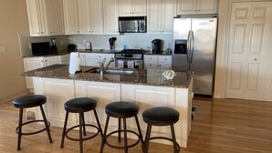 Een moderne keuken met witte kastjes, een roestvrijstalen koelkast, een magnetron en een granieten aanrecht. Er staan vier stoelen bij het eiland.