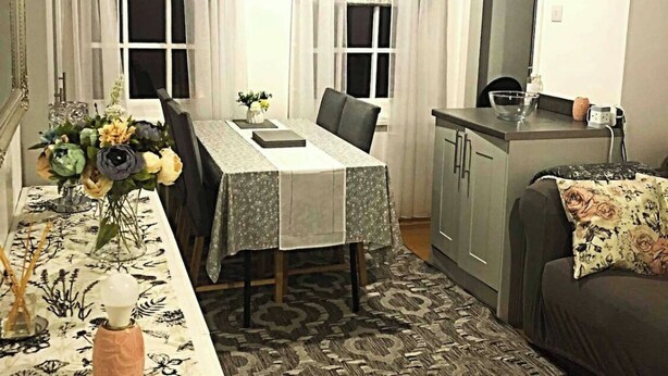 Un coin repas avec une table dressée pour deux personnes, un canapé, un espace cuisine et un tapis à motifs.
