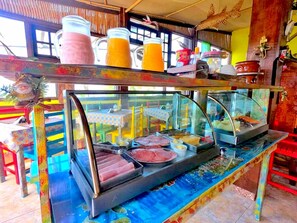 Um balcão de café da manhã com estrutura de vidro, diversas opções de alimentos e decoração colorida.
