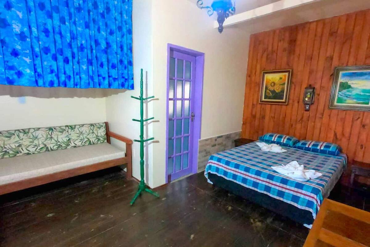 A room with a wooden floor, a purple door, a bed with a checkered bedspread, a wooden bench, and a green plant stand.