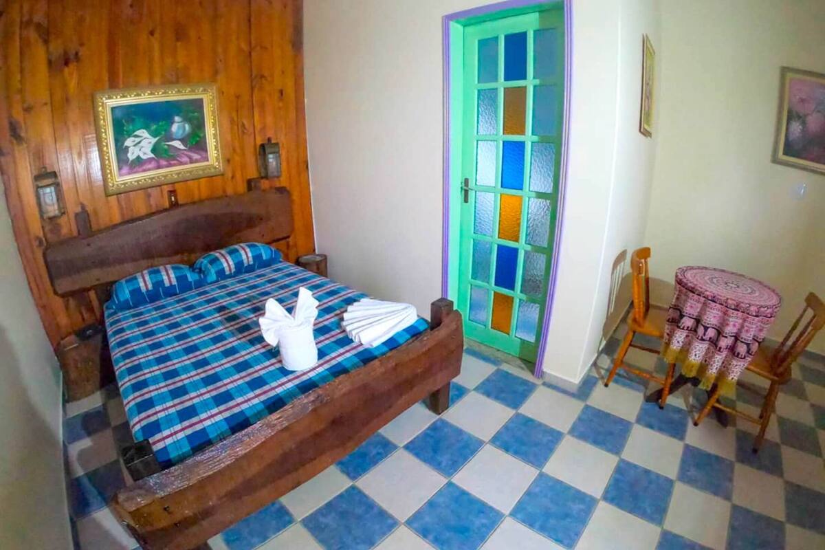 A room with a wooden bed, checkered floor, a green door, and a small round table with a patterned tablecloth.