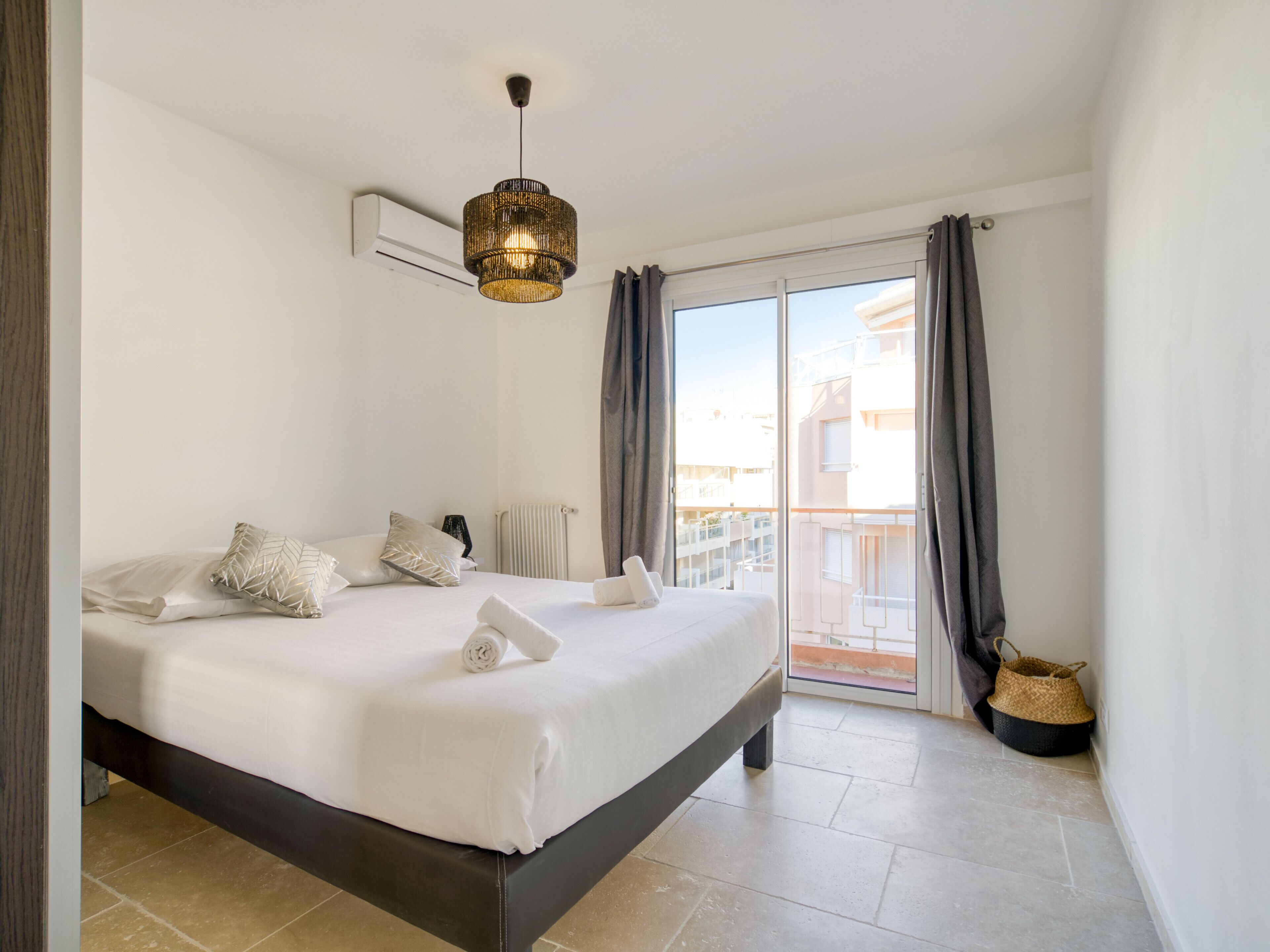 Una habitación de hotel con una cama grande, una lámpara en el techo, un radiador, un balcón con cortinas y una cesta tejida.
