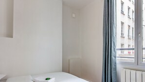 A hotel room with a bed, a window with curtains, a radiator, and a view of buildings outside. - Appartement des Bergères - Welkeys (Paris)
