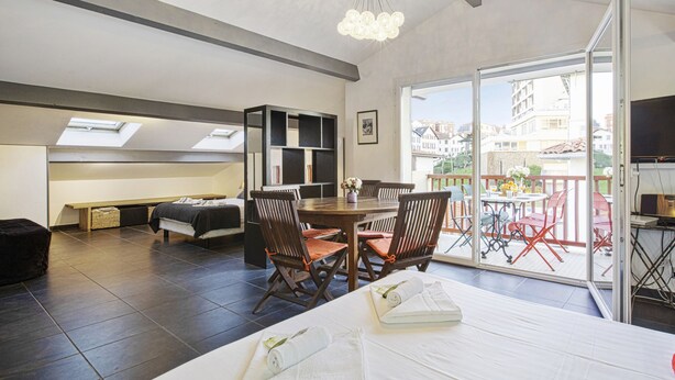 Une chambre moderne avec un grand lit, une table à manger avec des chaises, une bibliothèque intégrée et un balcon offrant une vue sur les bâtiments.