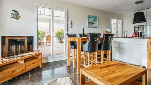 A modern living room with a TV, wooden furniture, a dining area with chairs, and a kitchen with a black lamp. - Appartement des Asphodèles - Welkeys (La Teste-de-Buch)