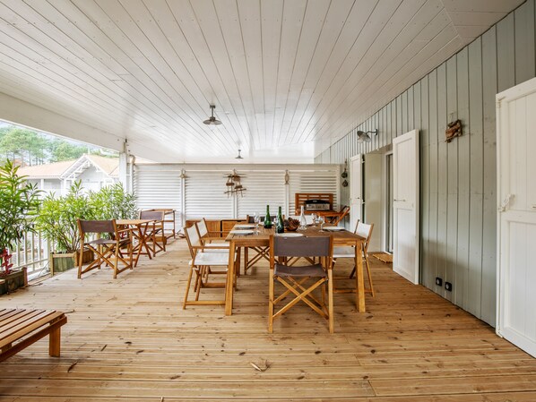 Une terrasse couverte avec un plancher en bois, une table à manger entourée de chaises, et une vue sur une maison en arrière-plan.