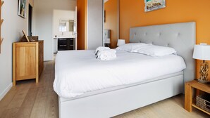 A modern hotel room with a large bed, a wooden dresser, a bathroom with a mirror, and a painting of a beach scene. - Appartement des Asphodèles - Welkeys (La Teste-de-Buch)