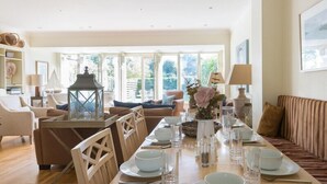 Un coin repas avec une longue table dressée pour quatre personnes, entourée de chaises en bois. On aperçoit un canapé et un banc rembourré au fond.