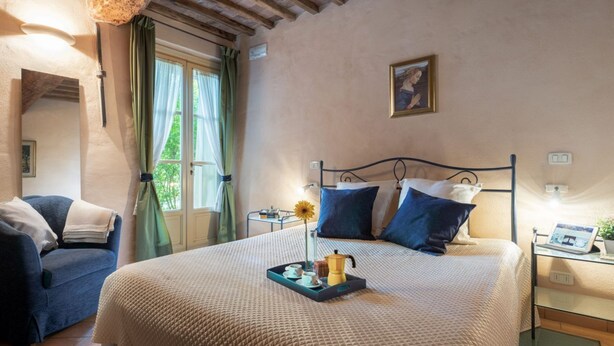 Een slaapkamer met een bed, nachtkastje met lamp, stoel en raam met gordijnen.