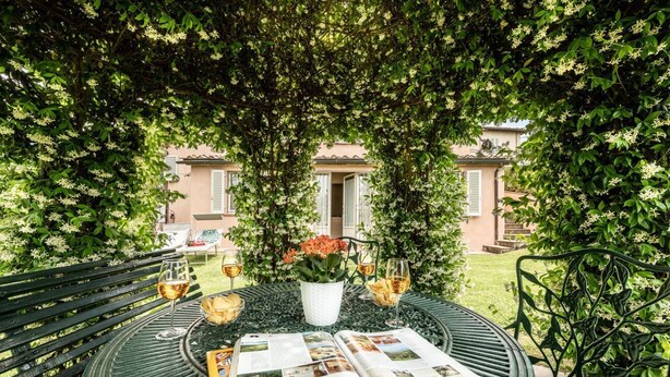 Una mesa metálica redonda con un libro y copas de vino, rodeada de vegetación exuberante y un edificio al fondo.