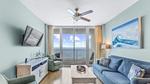 A spacious living room with a balcony, a ceiling fan, a flat-screen TV, and a painting.