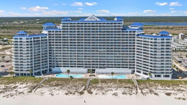 A large, multi-story hotel with a modern design, featuring a central courtyard with a pool and a beach area in the foreground.