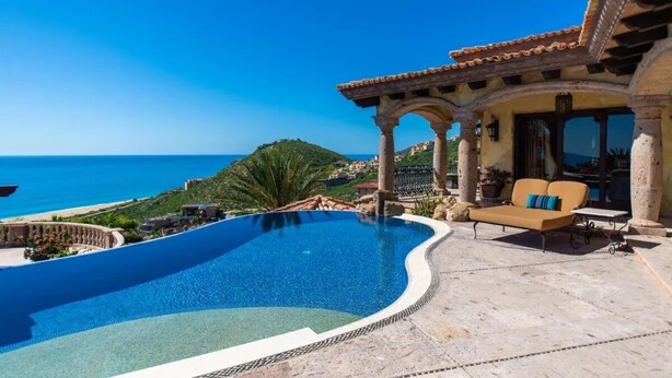 A pool with a view of the sea, a patio with a sofa, and a building with arches.