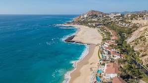 Vista aérea de una zona costera con playa arenosa, un rompeolas rocoso y edificios residenciales con techos de tejas rojas.