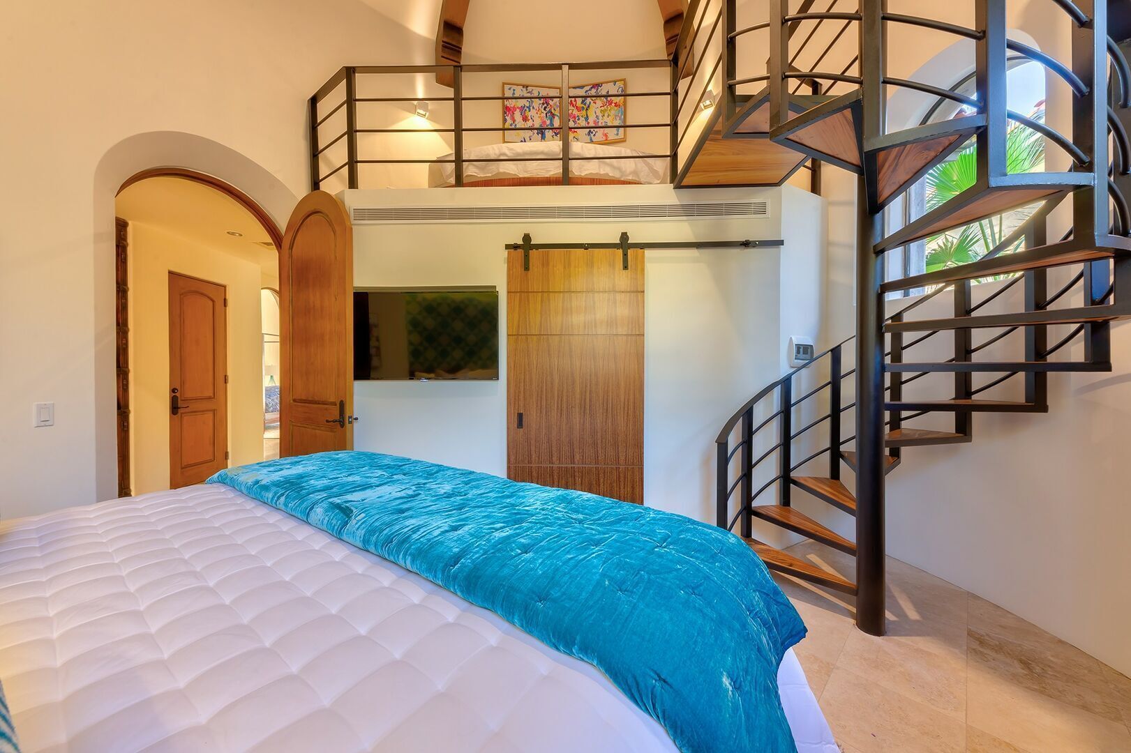A bedroom with a bed, a wooden wardrobe, a built-in wardrobe, a staircase, and a window with a view of greenery.