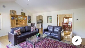 A spacious living room with two leather sofas, a glass coffee table, and a patterned rug. There is a wooden bookshelf with books and paintings, and a kitchen area is visible in the background.
