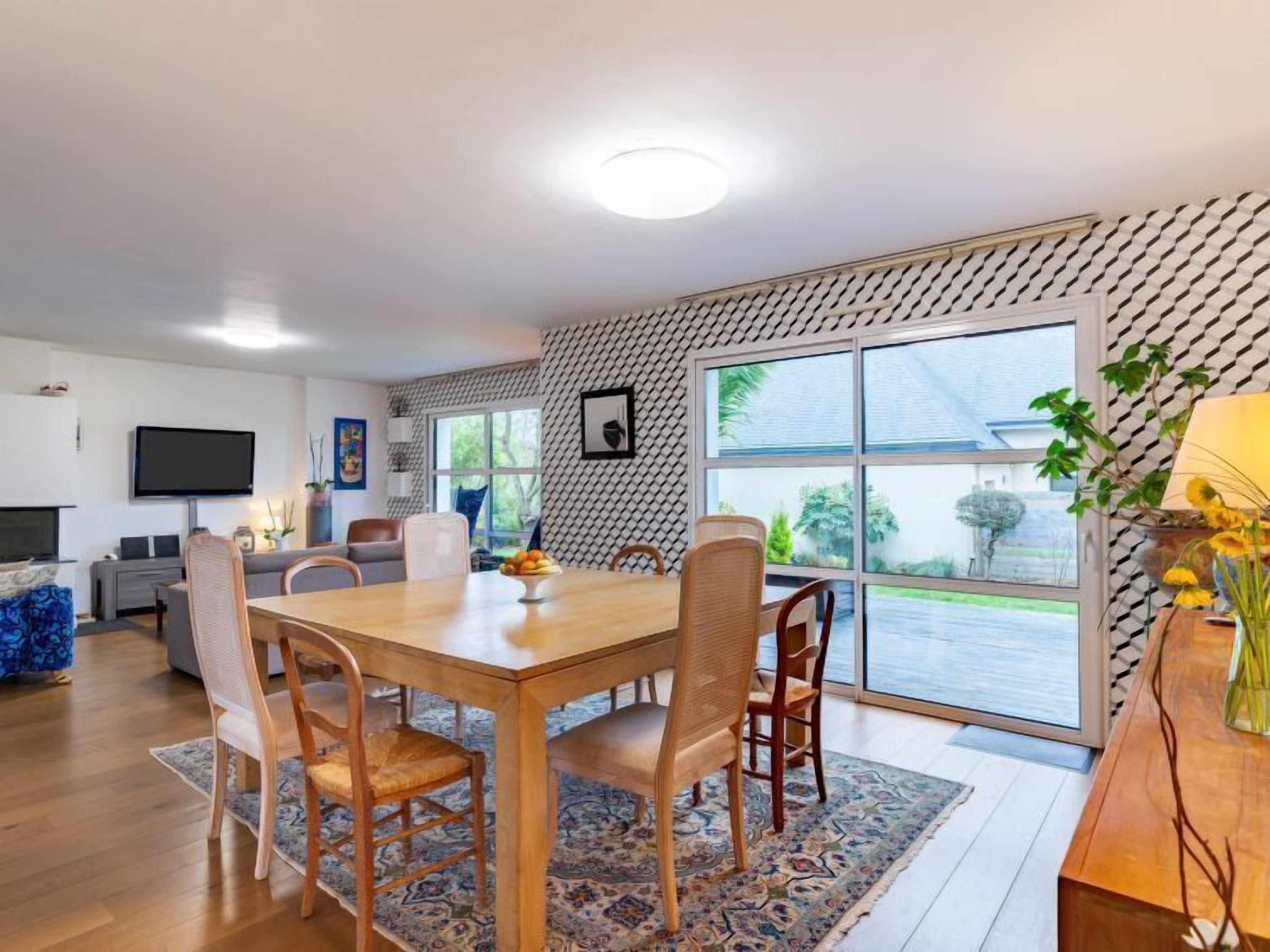 A modern living room with a wooden dining table, comfortable seating, a flat-screen TV, and a large window offering a view of the outdoors.