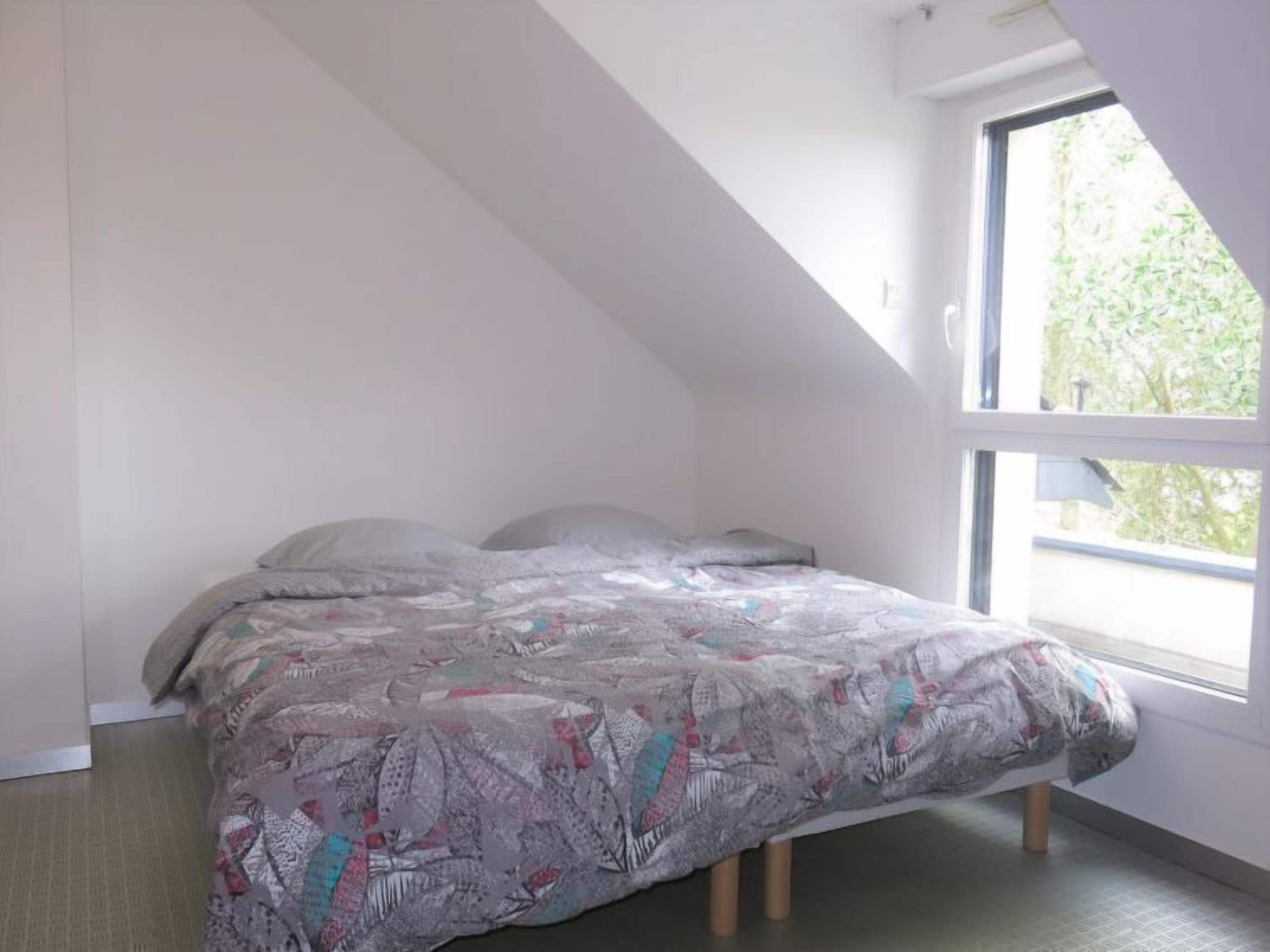 Een slaapkamer met een schuine kap, een bed met een bedspread met een patroon, en een raam met uitzicht op groen.