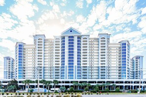 Stunning hotel with a central peaked roof and balconies on each floor, surrounded by lush palm trees and a covered walkway.