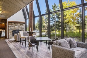 Un intérieur moderne avec une cheminée en pierre, un plafond en bois et de grandes fenêtres offrant une vue sur les arbres et une route.