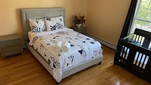 Ein ordentlich bezogenes Bett mit floralem Überwurf, ein Nachttisch mit Schublade, ein kleiner Tisch mit Blumenvase und ein Babybett in der Ecke.